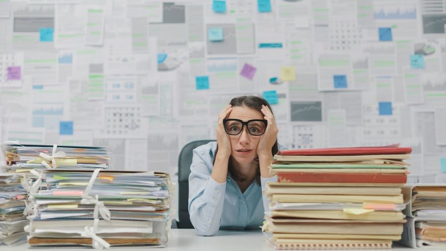 Una persona in ufficio circondata da pile di faldoni e documenti, con le mani sulla testa
